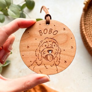 May include: A round wooden ornament with a laser-engraved image of a dog with the name "BOBO" written above it. The ornament is surrounded by small stars.
