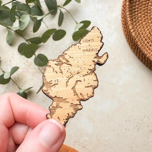 May include: A wooden map of Lewis and Harris, with the names etched into the wood. The map is light brown with darker brown edges, and is held in a hand. The background includes green leaves and a woven basket.