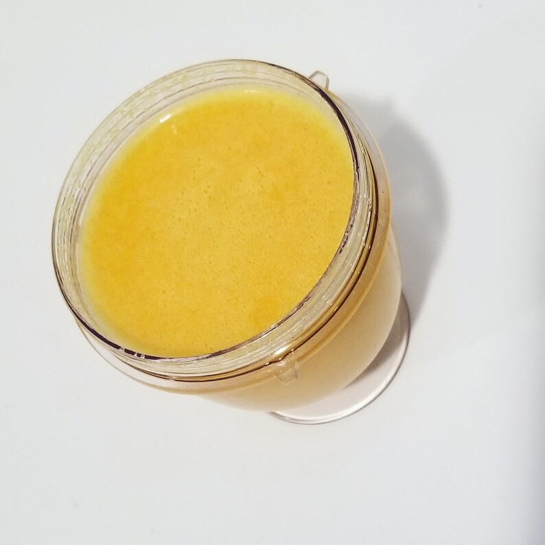 May include: A clear glass container filled with a thick, yellow-orange liquid. The container has a rounded base and a handle. The liquid appears smooth and has a slight foam on the surface. The container is set against a white background.