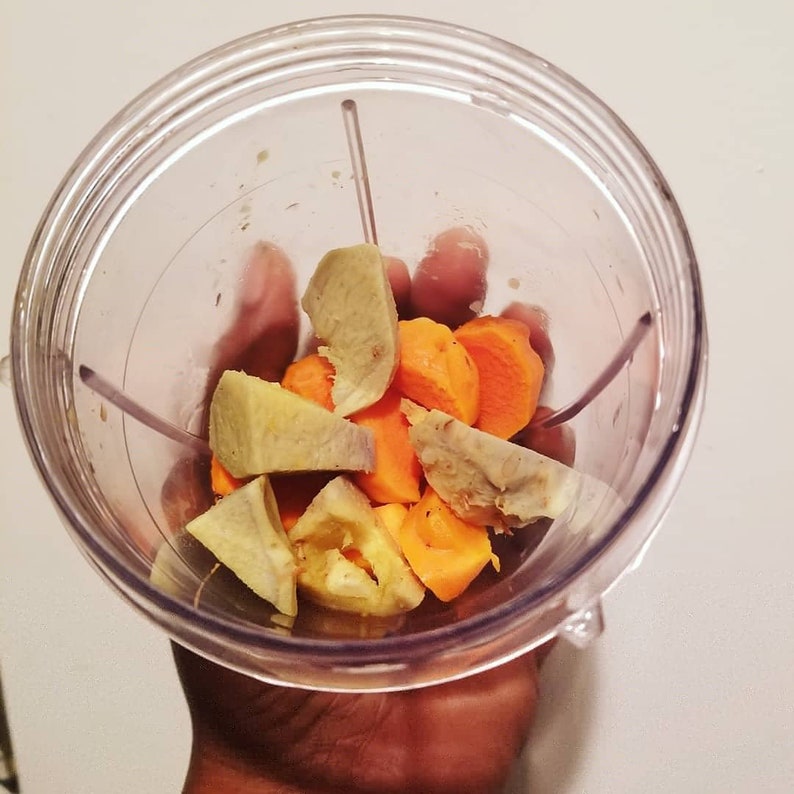 May include: A clear plastic blender cup filled with chunks of orange carrots and light brown ginger. The cup is held by a hand, with a white background.