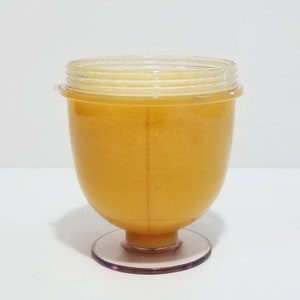 May include: A clear, plastic container with a light orange liquid inside. The container has a rounded body, a clear lid, and a pink base. The container is sitting on a white surface.