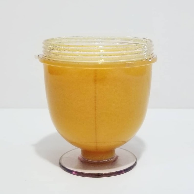 May include: A clear, plastic container with a light orange liquid inside. The container has a rounded body, a clear lid, and a pink base. The container is sitting on a white surface.