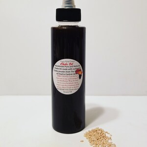 May include: A dark brown glass bottle with a black cap and a white label that reads "Chebe Oil Handmade Pure and Natural Chebe Oil made with authentic Chebe powder from The Republic of Chad in Central Africa". The bottle is sitting on a white surface with a small pile of light brown sesame seeds in front of it.