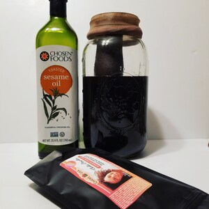 May include: A glass jar with a brown lid filled with a dark liquid. A bottle of Chosen Foods Toasted Sesame Oil is next to the jar. A black pouch with a pink label that reads "Chebe USA Chebe Powder Authentic From Chad" is in the foreground.