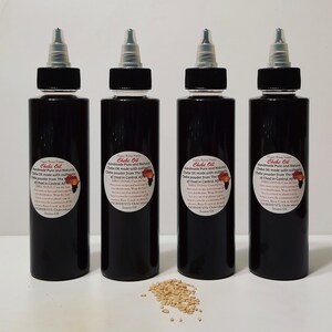 May include: Four dark brown glass bottles with black caps and labels that read "Chebe Oil Handmade Pure and Natural Chebe Oil made with authentic Chebe powder from The Republic of Chad in Central Africa". The bottles are arranged in a row on a white surface. A small pile of light brown seeds is in front of the bottles.