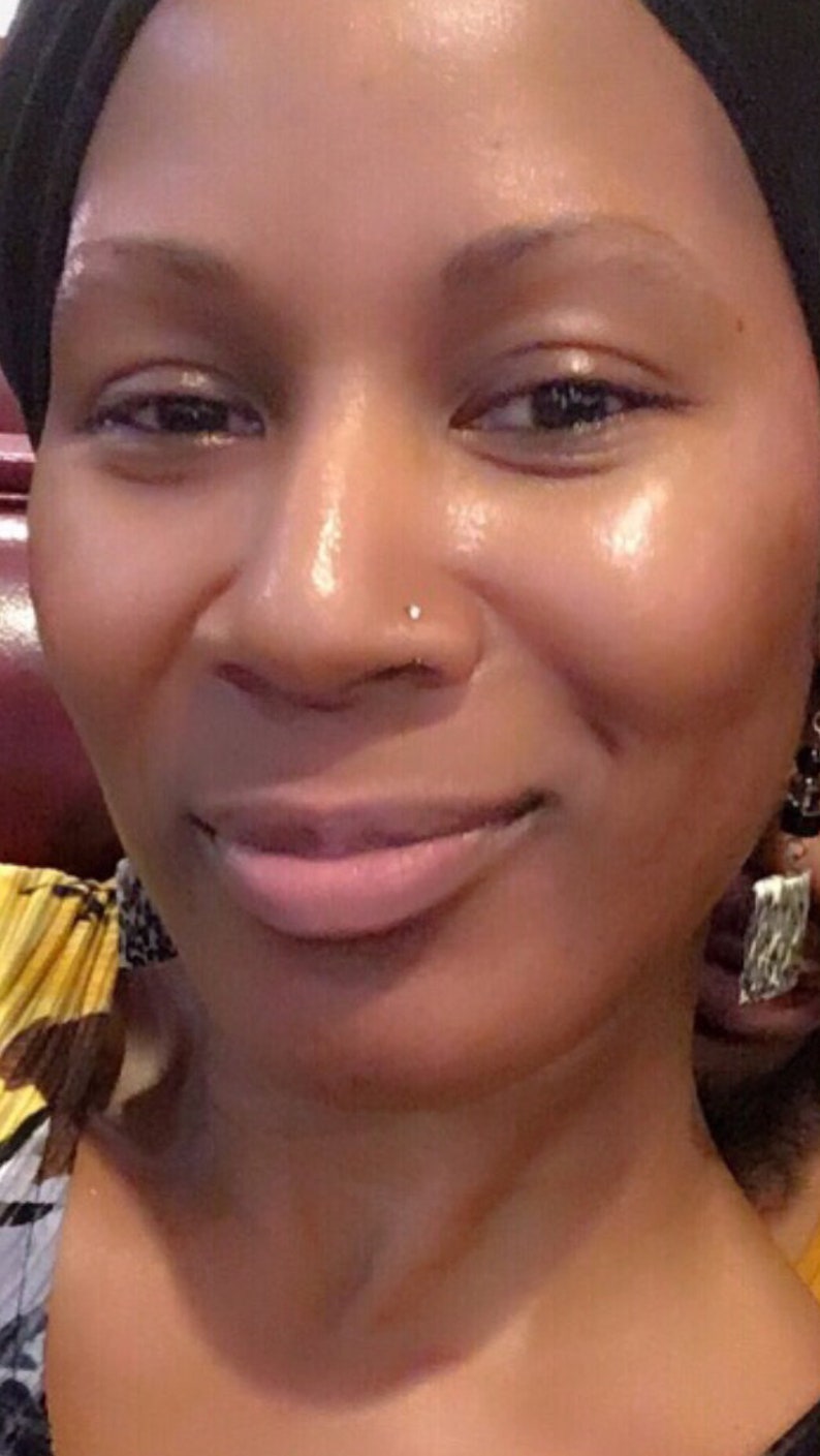 May include: A close-up portrait of a person with brown skin, wearing a black head covering. The person has a nose piercing and is smiling. The background is blurred, with hints of yellow and black patterns.