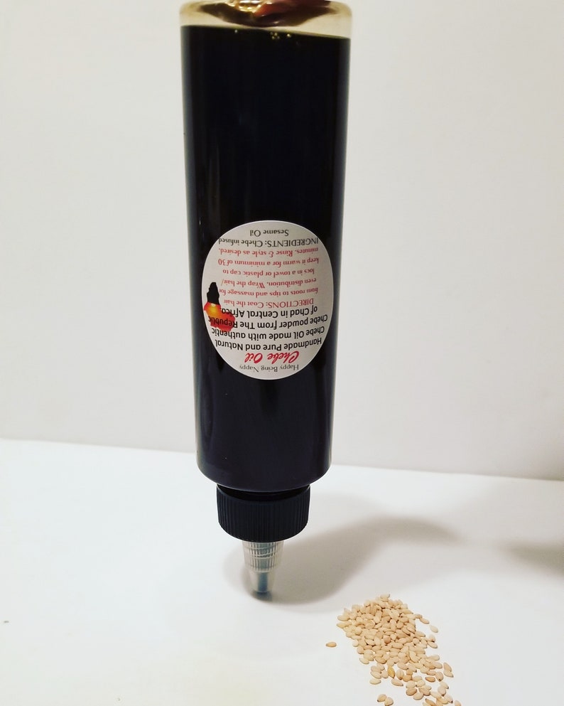 May include: A black bottle of Chebe Oil with a white label that reads "Chebe Oil Handmade Pure and Natural Chebe Oil made with authentic Chebe powder from The Republik DIRECTIONS Coathe hair Forestops and massage Prediaribution. Wrap the har locs in a towel or plastic cap to keep it was for a minimum of 30 mintes. Rise & style as desired. INGREDIENTS: Chebe infused. Sesame Oil". A small pile of sesame seeds is in the foreground.