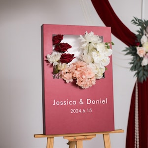 May include: A burgundy wedding sign with a white frame and floral arrangement. The sign reads "Jessica & Daniel 2024.6.15".