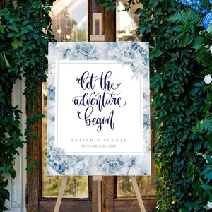 May include: A wedding welcome sign with a blue watercolor background and white text that reads "Let the adventure begin" with the couple's names and wedding date below. The sign is on a wooden easel in front of a doorway decorated with blue and white flowers.