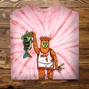 May include: A tie-dye t-shirt featuring a cartoon mascot wearing a basketball jersey with the text "HEAT 1" and holding a fish. The mascot is fishing with a rod and reel. The background is a pink and white tie-dye pattern.