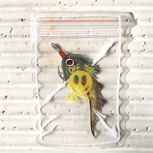 May include: A clear plastic bag with a zipper closure contains a yellow and black fishing lure with a smiley face and the text "Off the Grid".
