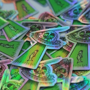 May include: A pile of holographic stickers featuring a green alien spaceship with two aliens inside. The spaceship is flying over a green fish. The stickers have the text "Off the Grid John" on them.