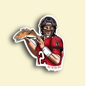 May include: A digital illustration of a football player wearing a red jersey with a black number "2" on the front, a black helmet, and a white wristband. The player is holding a red fish with a white belly. The text "OFF THE GRID JOHN" is below the player.