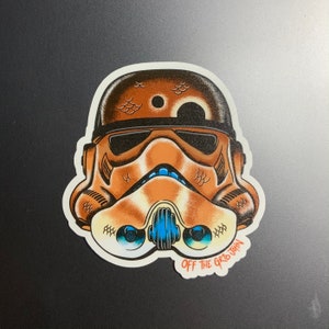 May include: A brown and white Star Wars Stormtrooper helmet sticker with a blue and white face mask. The helmet has a black visor and a brown and white pattern on the top. The sticker has the text "OFF THE GRID TOWN" in red at the bottom.