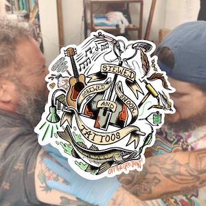 May include: A colorful tattoo design featuring a fishing lure, a guitar, a fish, and the words "Stewed and Snook Tattoos" with the tagline "Off the Grid John".