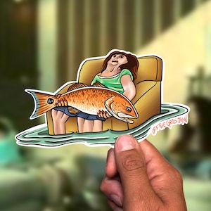 May include: A sticker featuring a cartoon illustration of a person sitting in a yellow armchair holding a large red fish. The sticker has a white border and the text "OFF THE GRID JOHN" in black lettering.