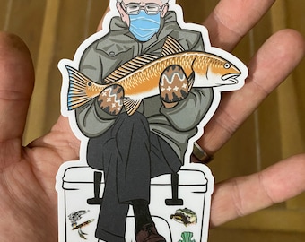 May include: A sticker featuring a cartoon illustration of a man wearing a face mask and mittens, holding a fish. The man is seated on a cooler with fishing-themed stickers. The text "OFF THE GRID JOHN" is at the bottom.
