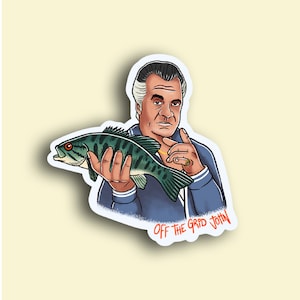 May include: A sticker featuring a cartoon illustration of a man holding a fish. The man is wearing a blue jacket and has a serious expression. The text "OFF THE GRID JOHN" is written in orange below the man.