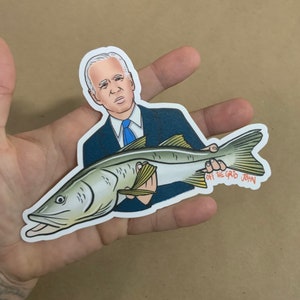 May include: A sticker featuring a cartoon illustration of a person holding a fish. The person is wearing a blue suit and a blue tie. The fish is a snook, a type of fish commonly found in Florida. The sticker has the text "Off the Grid John" on it.