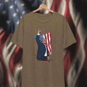 May include: A brown t-shirt with a graphic of a man holding a fishing rod and a US flag. The man is wearing a blue suit and has a raised fist. The text "We Are Winning" is printed below the graphic.