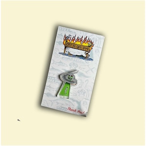 May include: A green and white enamel pin featuring a cartoon UFO with two aliens inside. The pin is on a white card with a cartoon image of a burning sign that reads "Off the Grid John".