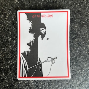 May include: Black and white sticker with a red border. The sticker features a cartoon illustration of a man holding a fishing rod. The text "Off the Grid John" is written in black at the top of the sticker.