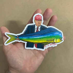 May include: A sticker featuring a cartoon illustration of a man wearing a red "Make Fishing Great Again" hat holding a large yellow, blue, and green fish. The text "Off the Grid John" is printed below the fish.