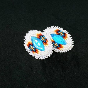 May include: A pair of oval-shaped beaded earrings featuring a central marquise-cut bright blue crystal. The border consists of white seed beads, with inner rows of orange, red, black, blue, and metallic gold beads surrounding the blue crystal center.