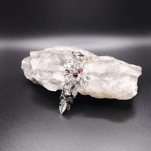 Op de afbeelding: Een zilverkleurige metalen sleutelhanger met een karabijnsluiting, meerdere ringetjes en bungelende bedels. De bedels bestaan uit kleine rode en grijze kralen, samen met gedetailleerde metalen hangers in de vorm van bladeren.
