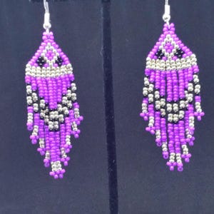 May include: A pair of dangle earrings crafted from purple, silver, and black seed beads. The earrings feature a geometric pattern at the top with a fringe of beaded strands hanging below, suspended from silver-toned metal earring hooks.