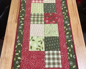 May include: A red and green patchwork table runner with a white snowflake and holly berry pattern. The runner has a green and white border with a red trim.
