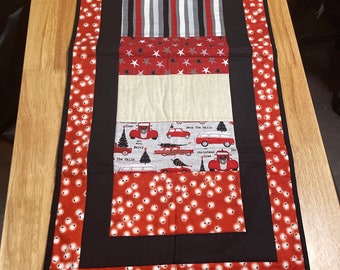 May include: A red, black, and white Christmas table runner with a repeating pattern of white lights on a red background. The runner features a variety of Christmas-themed designs, including red trucks, stars, and the words "Deck the Halls" and "Merry Christmas".