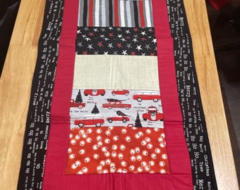 May include: Red, black, and white Christmas table runner with a festive design featuring ornaments, stars, and vintage cars. The runner has a black border with the text "Merry Christmas Merry" repeated around the edge.