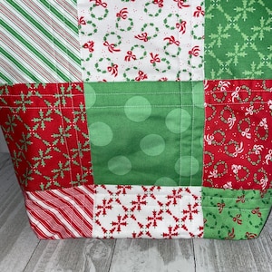 May include: A patchwork tote bag with a Christmas theme. The bag is made of different fabrics with red, green, and white patterns, including snowflakes, candy canes, and wreaths.