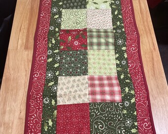May include: A red and green patchwork table runner with a white snowflake and holly berry design. The runner has a red border with a green and white floral pattern.