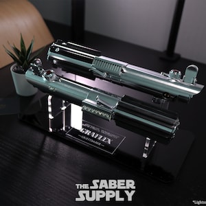 Puede incluir: Un soporte de exhibici&oacute;n de sable de luz negro y plateado con el texto "The Saber Supply" y "Graflex Lightsaber" en la base. *Sable de luz no incluido*.