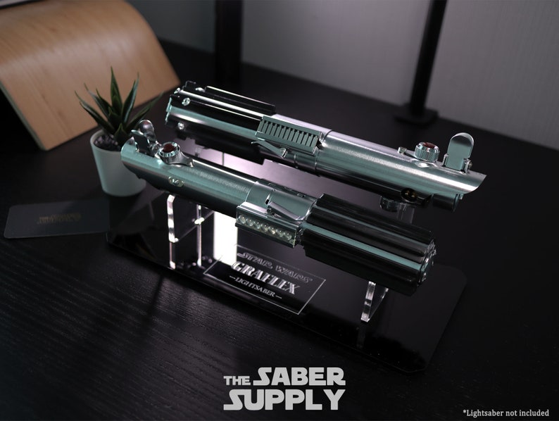 Puede incluir: Un soporte de exhibici&oacute;n de sable de luz negro y plateado con el texto "The Saber Supply" y "Graflex Lightsaber" en la base. *Sable de luz no incluido*.