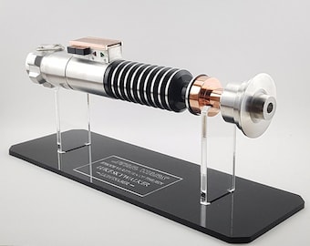 May include: A silver and black lightsaber replica with a copper accent on a black display stand. The stand has a plaque that reads "STAR WARS EPISODE V: THE EMPIRE STRIKES BACK - LUKE SKYWALKER - LIGHTSABER".