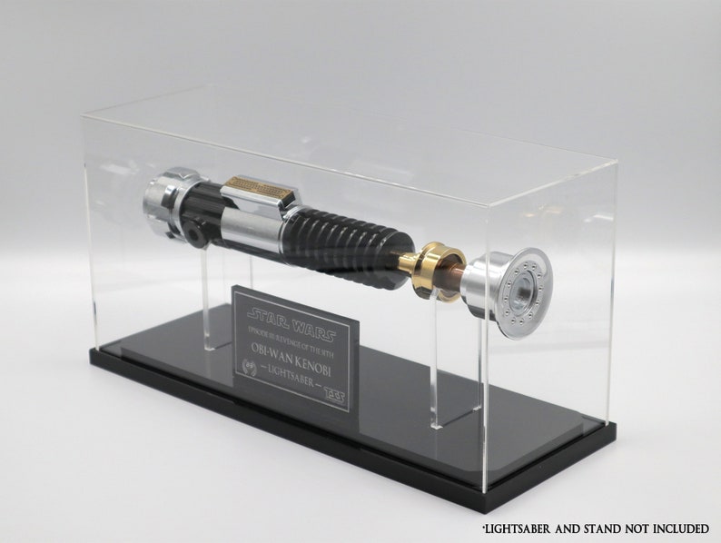 Puede incluir: Una vitrina de acr&iacute;lico transparente con una base negra, que muestra una r&eacute;plica de sable l&aacute;ser plateado y negro. La vitrina tiene una placa negra con el texto "STAR WARS EPISODE III REVENGE OF THE SITH OBI-WAN KENOBI LIGHTSABER - 1:1"