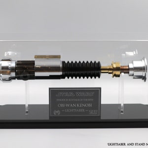 Puede incluir: Una r&eacute;plica de sable de luz negro y plateado de Star Wars Episodio III: La venganza de los Sith, expuesta en una vitrina de acr&iacute;lico transparente con una base negra. La vitrina presenta una placa con el texto "STAR WARS EPISODE III REVENGE OF THE SITH OBI-WAN KENOBI - LIGHTSABER - 833".