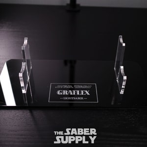 Puede incluir: Un expositor de acr&iacute;lico negro con dos soportes de acr&iacute;lico transparente para sables de luz. El expositor presenta el texto "STAR WARS GRAFLEX - LIGHTSABER -" en letras blancas.