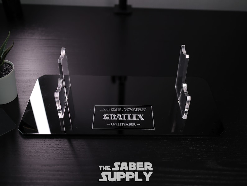 Puede incluir: Un expositor de acr&iacute;lico negro con dos soportes de acr&iacute;lico transparente para sables de luz. El expositor presenta el texto "STAR WARS GRAFLEX - LIGHTSABER -" en letras blancas.