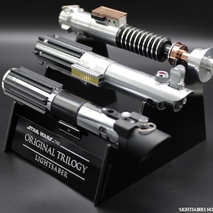 May include: A black display stand holding three lightsabers. The stand has the text "STAR WARS - THE ORIGINAL TRILOGY LIGHTSABER". The lightsabers are silver and black, with detailed designs. The lightsabers are not included.