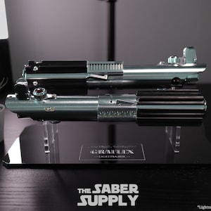 Puede incluir: Dos r&eacute;plicas de sables de luz plateadas y negras en un soporte de exhibici&oacute;n negro con una base de acr&iacute;lico transparente. El soporte tiene una placa negra con el texto "GRAF LEX - LIGHTSABER -" en ella. *Sable de luz no incluido*.