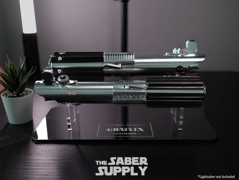 Puede incluir: Dos r&eacute;plicas de sables de luz plateadas y negras en un soporte de exhibici&oacute;n negro con una base de acr&iacute;lico transparente. El soporte tiene una placa negra con el texto "GRAF LEX - LIGHTSABER -" en ella. *Sable de luz no incluido*.