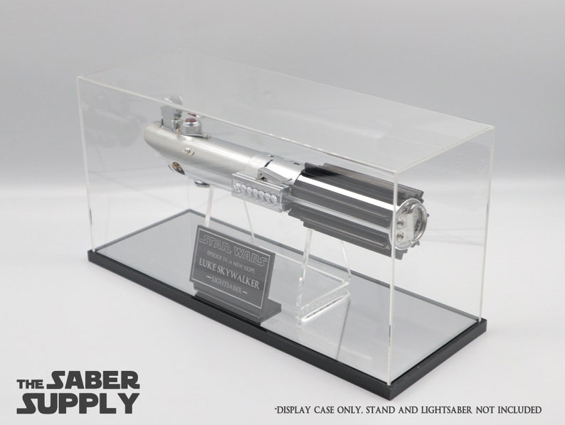 K&ouml;nnte beinhalten: Eine transparente Acryl-Vitrine mit schwarzem Sockel, die eine silberne Lichtschwert-Replik zeigt. Die Vitrine enth&auml;lt eine schwarze Plakette mit dem Text "STAR WARS", "LUKE SKYWALKER" und "LIGHTSABER".