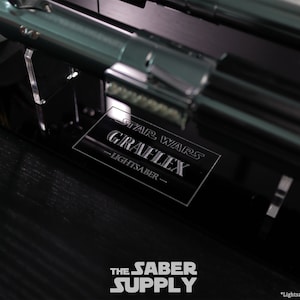 Puede incluir: Un soporte de exhibici&oacute;n negro con una placa plateada que dice "Star Wars Graflex Lightsaber". El soporte est&aacute; sobre una superficie negra. *Sable de luz no incluido*.
