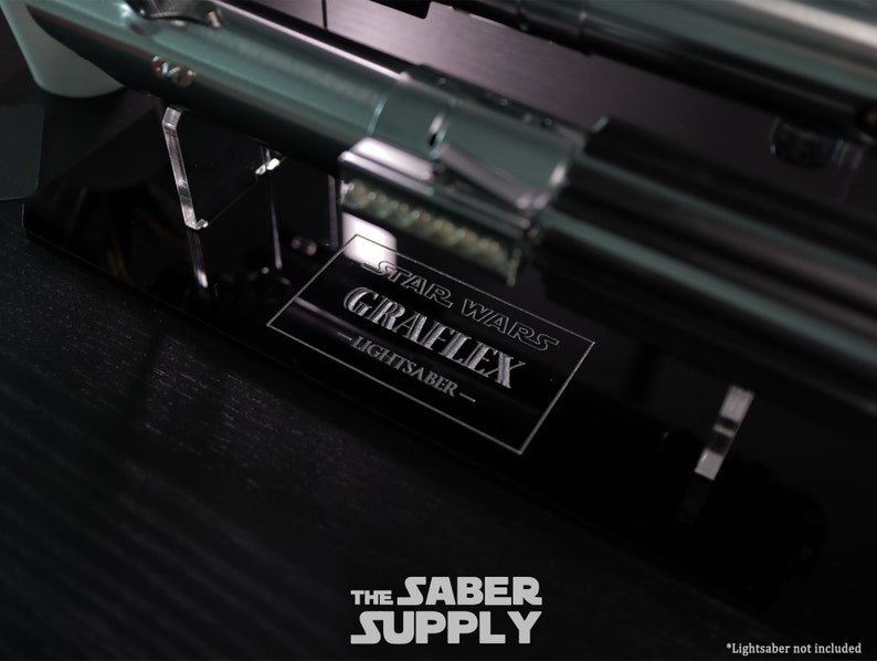 Puede incluir: Un soporte de exhibici&oacute;n negro con una placa plateada que dice "Star Wars Graflex Lightsaber". El soporte est&aacute; sobre una superficie negra. *Sable de luz no incluido*.