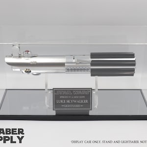 K&ouml;nnte beinhalten: Eine transparente Acryl-Vitrine mit schwarzem Sockel und einem verspiegelten Boden. In der Vitrine befindet sich eine silberne Lichtschwert-Replik mit schwarzem Griff. Die Vitrine hat eine schwarze Plakette mit dem Text "STAR WARS EPISODE IV: EINE NEUE HOFFNUNG LUKE SKYWALKER -LICHT SCHWERT- ". Der Text "NUR VITRINE. ST&Auml;NDER UND LICHT SCHWERT NICHT INBEGRIFFEN" ist unter der Vitrine gedruckt.