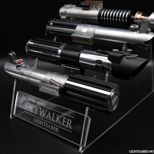 Puede incluir: Un expositor de acrílico transparente para sables de luz. El expositor tiene una base negra y un panel de acrílico transparente con el texto "Star Wars Skywalker Lightsaber" grabado en él. El expositor está diseñado para sostener cuatro sables de luz, y los sables de luz no están incluidos.